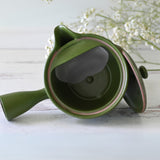 A Handcrafted Japanese Kyusu Teapot with Filters (380ml) in green rests on a white wooden surface, accompanied by small white flowers, capturing the elegance of Japanese ceramics.