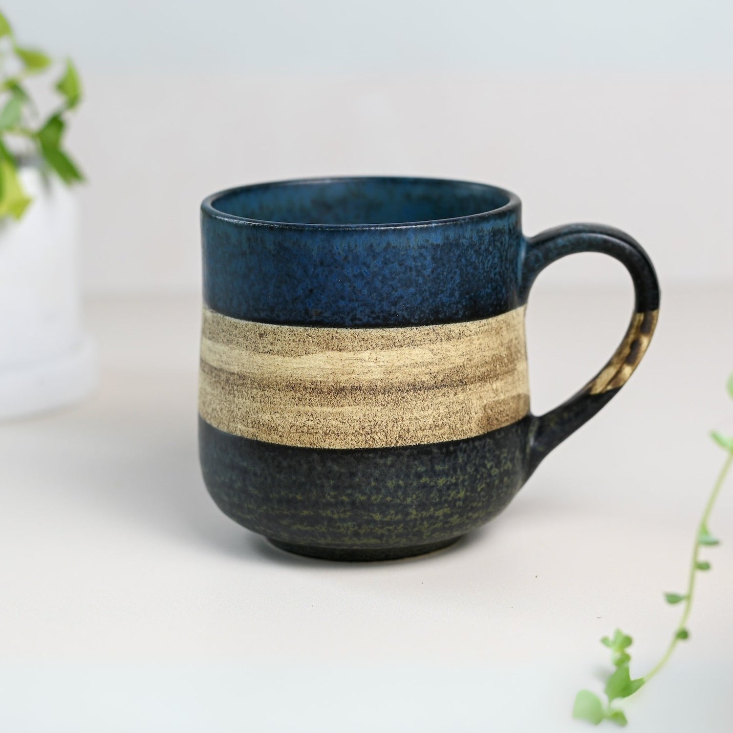 The Handcrafted Mino Ware Ceramic Mug from Japan, an 8 oz (240 ml) masterpiece, features blue, beige, and dark green bands on a light surface amidst lush greenery, evoking the elegance of Japanese pottery.