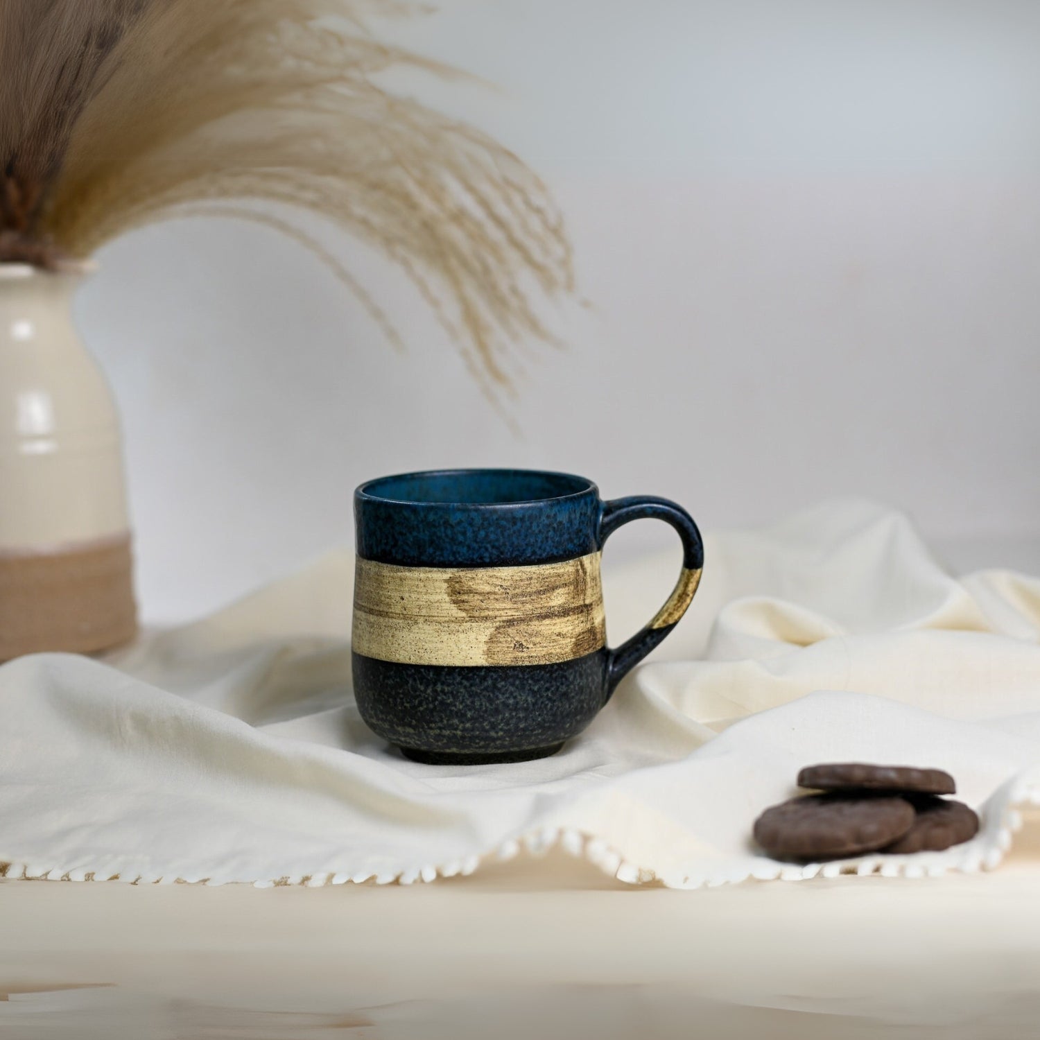 A Handcrafted Mino Ware Ceramic Mug from Japan, 8 oz (240 ml), with a blue hue and beige stripe, rests on a white cloth next to cookies and a vase of dry grass.