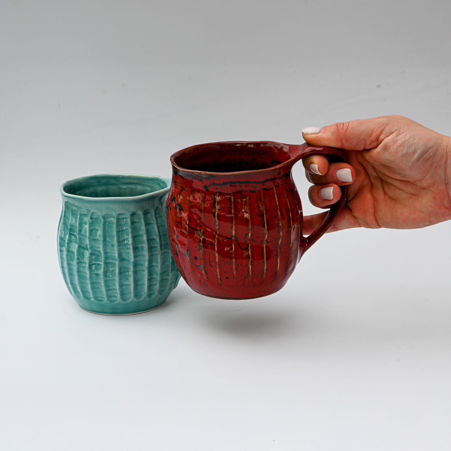 A hand holds a red mug next to the Handmade Big Blue Ceramic Tea or Coffee Mug from Japan (18oz), both set against a plain white background.