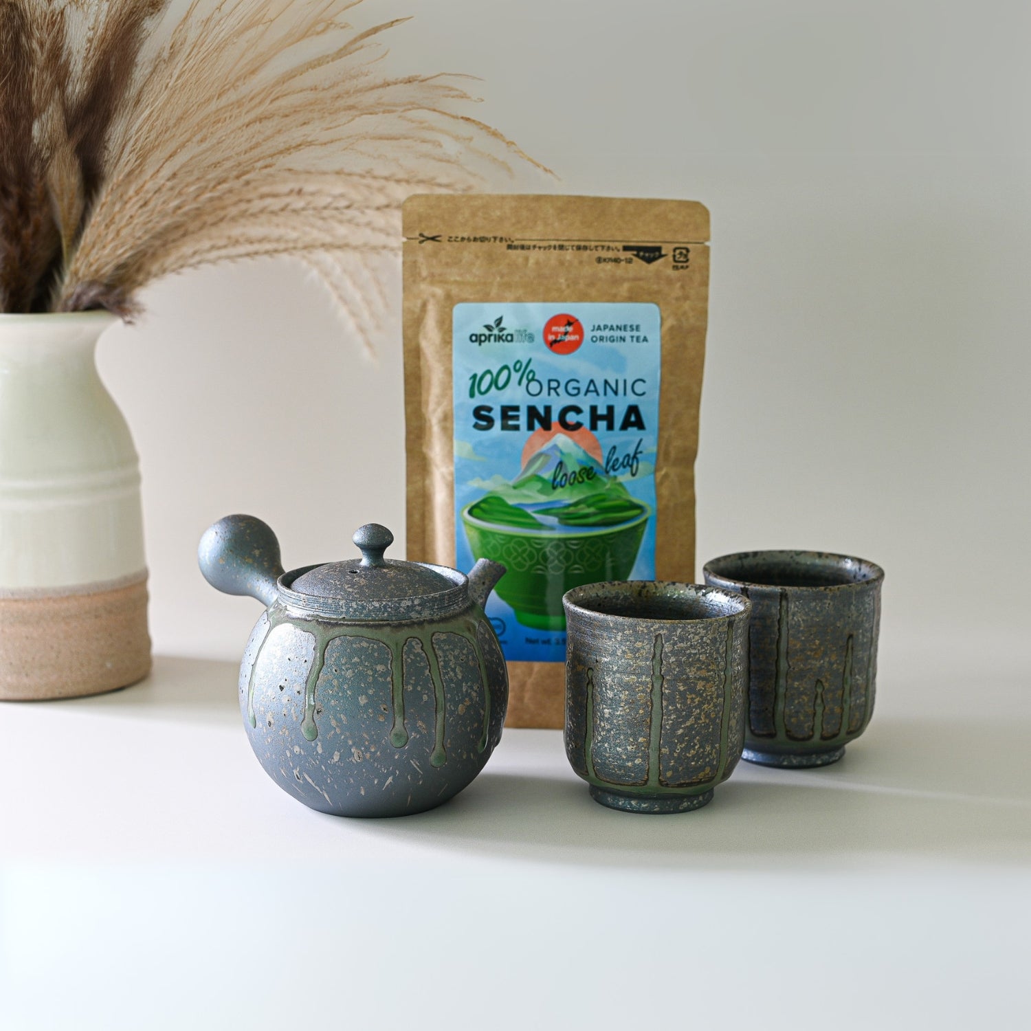 The Japanese Kyusu Set, featuring a 230ml teapot with filters and two cups, rests on a white surface alongside organic sencha tea and a pampas grass vase.