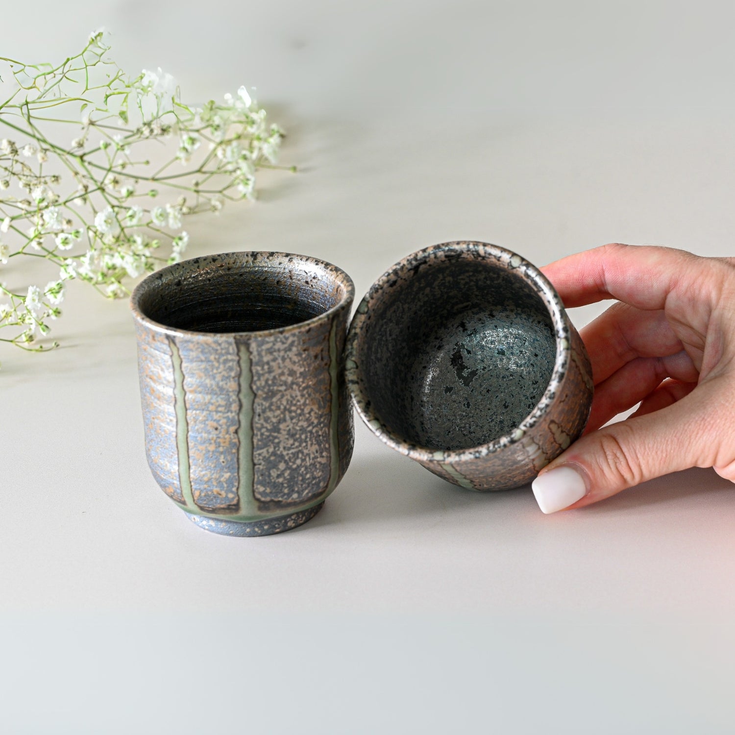 Holding a cup from the Japanese Kyusu Set - Teapot with Filters (230ml) and Two Cups, with an identical one beside it; baby's breath flowers accentuate the traditional artistry of Tokoname ceramics.