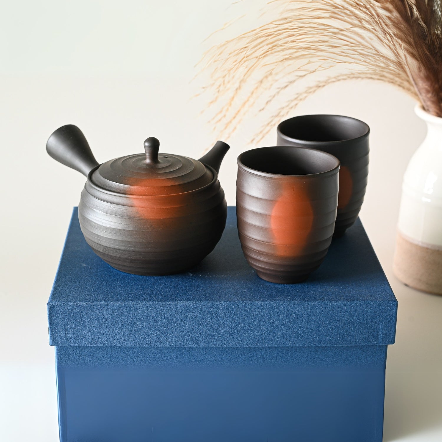 A charming display features a Japanese Kyusu Set with a 270ml teapot and two cups, elegantly placed on a blue box beside a vase of dried grasses.