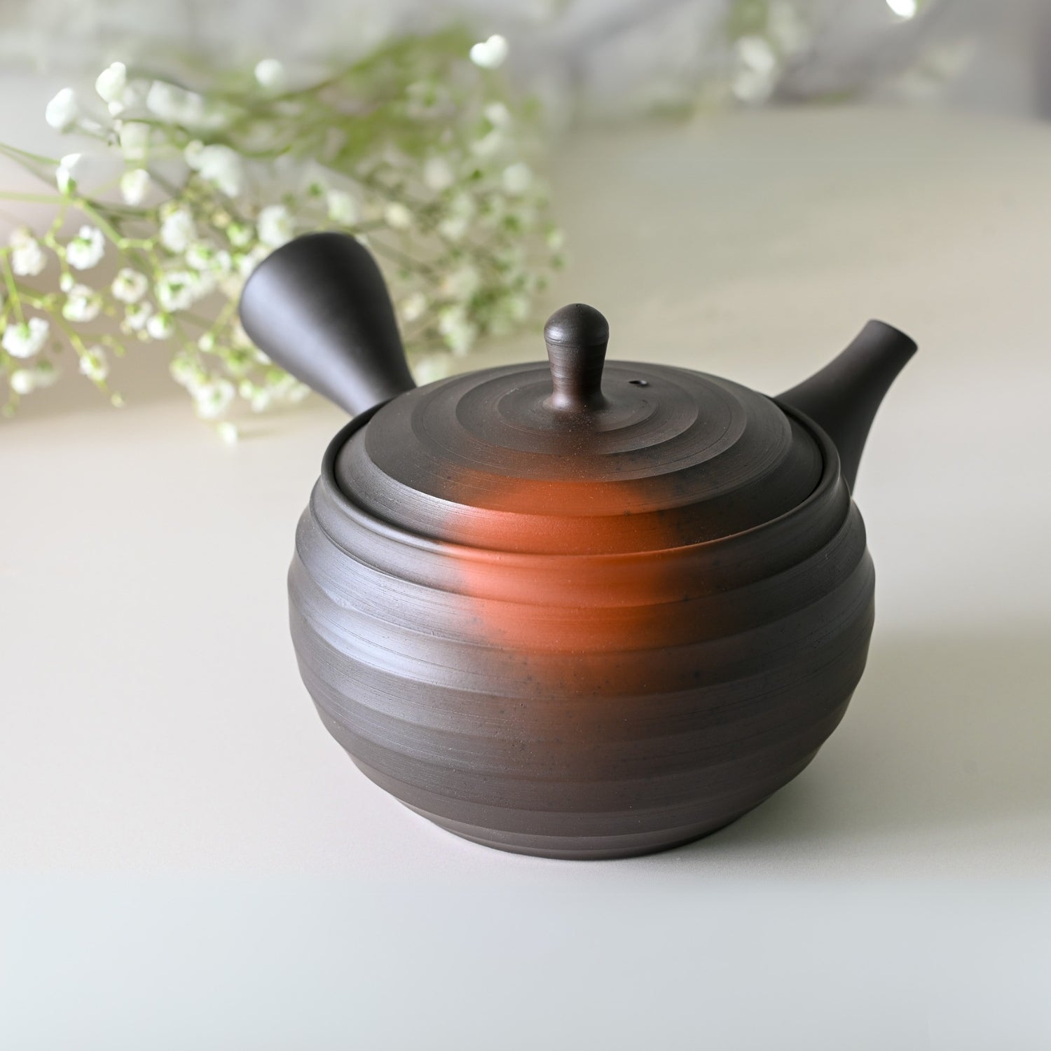 A Japanese Kyusu teapot (270ml), part of a set with filters and two cups, featuring a dark hue with a reddish tint, is elegantly surrounded by delicate white flowers on a smooth surface.