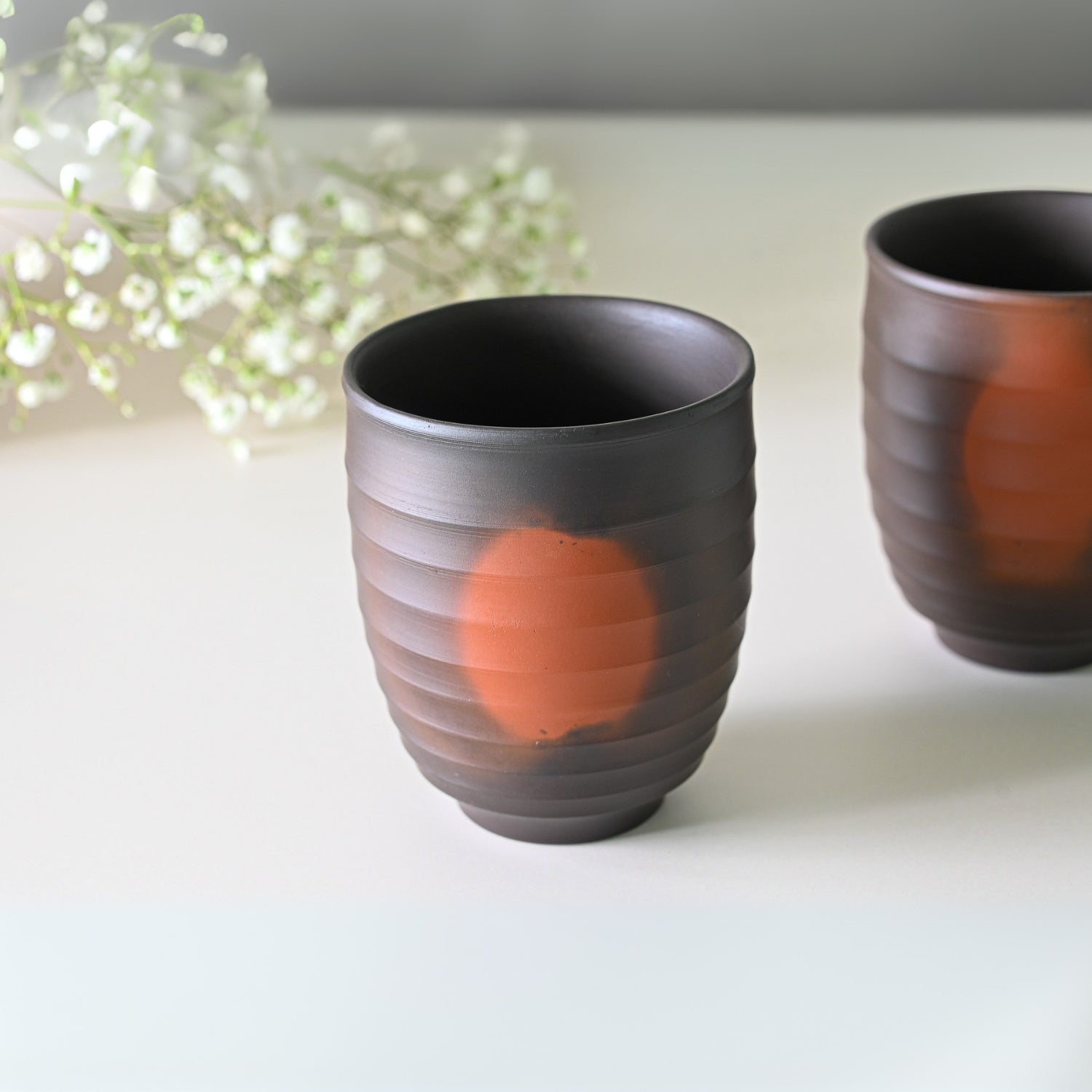 Two cups from the Japanese Kyusu Set, featuring a ribbed design and orange accents reminiscent of Yunomi Teacups, are placed on a white surface adorned with small white flowers.