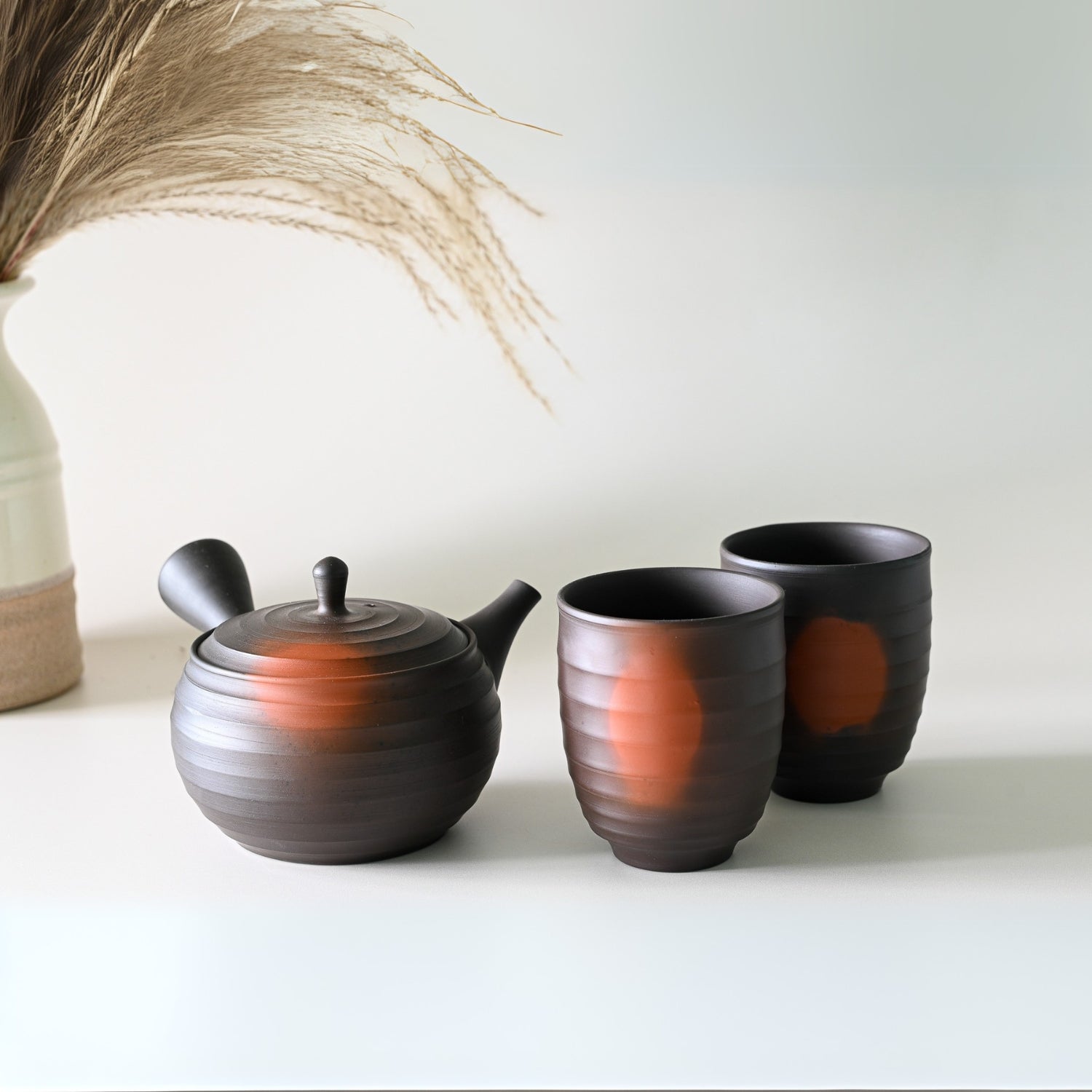 A Japanese Kyusu Set with a black ceramic teapot (270ml) and two Yunomi teacups featuring brown accents sits on a light surface alongside a vase of dried grass, echoing the elegance of Tokoname Ware style.