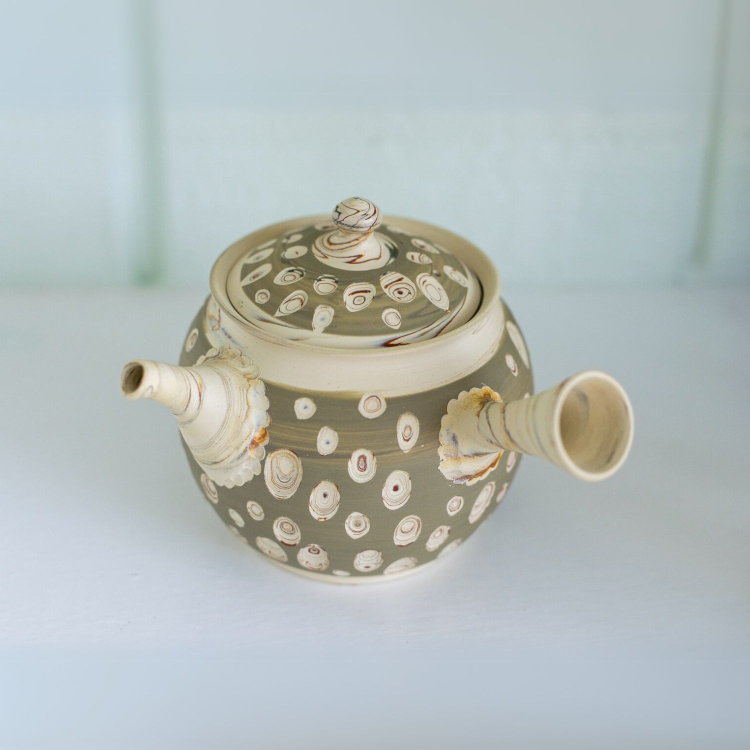 The Japanese Kyusu Set includes a 370ml teapot crafted from Tokoname clay with unique spout and lid, adorned with circle patterns on a light-colored background. It's perfect for brewing green tea and enhancing your tea-time experience.