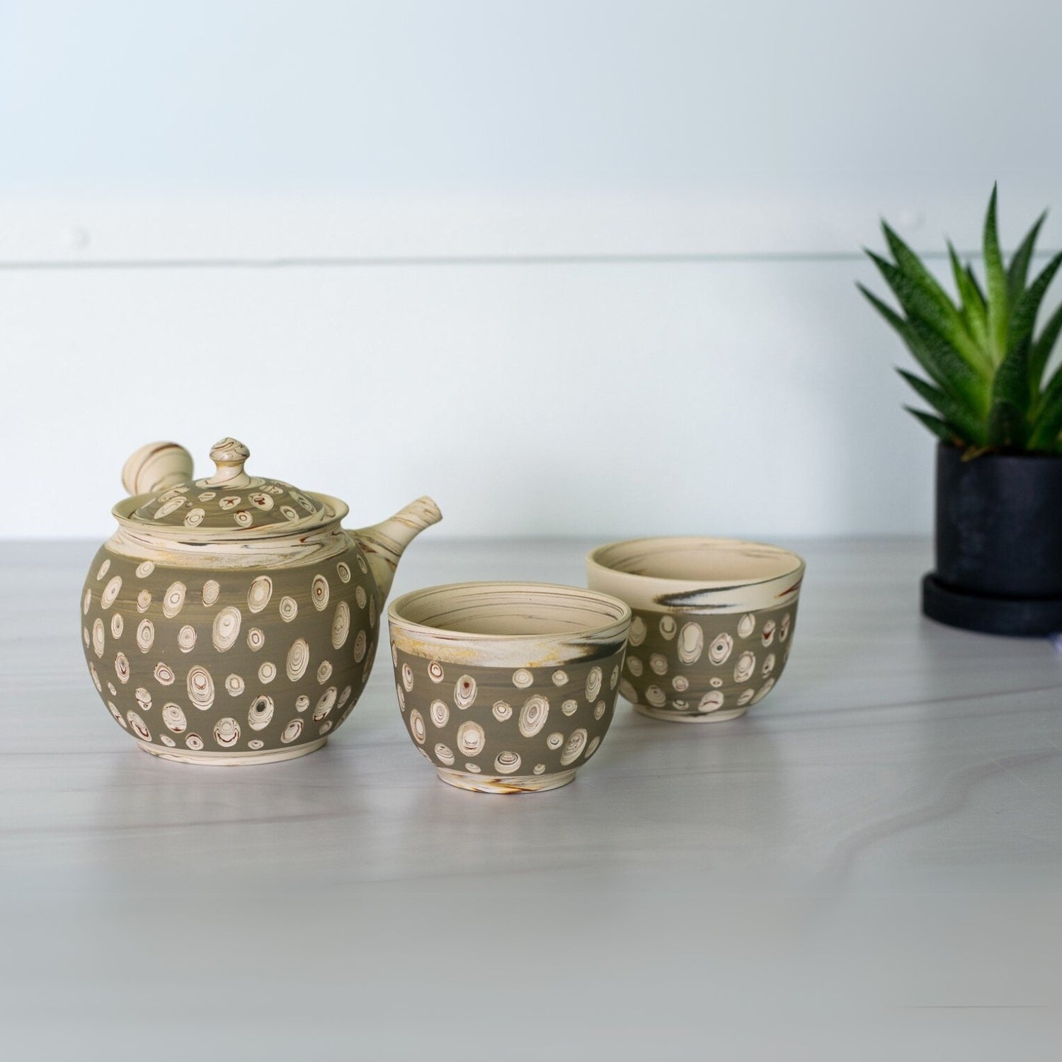 The Japanese Kyusu Set with a teapot (370ml) and two cups on a white table, accompanied by a potted succulent, invites you to enjoy a soothing green tea moment.