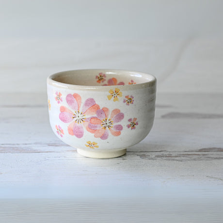 The Japanese Pink Sakura Ceramic Matcha Bowl, crafted in the traditional Minoyaki style with a pink and yellow floral design, rests on a light wooden surface.