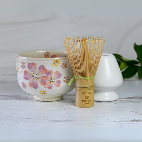 A Japanese Pink Sakura Ceramic Matcha Bowl, with pink flower motifs, is elegantly displayed with a bamboo matcha whisk and white ceramic holder on a light wooden surface.