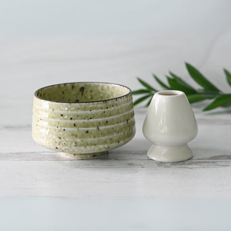 A Kaneni White Ceramic Matcha Bowl, crafted using Minoyaki techniques, and a small white vase sit on a light surface adorned with green leaves.