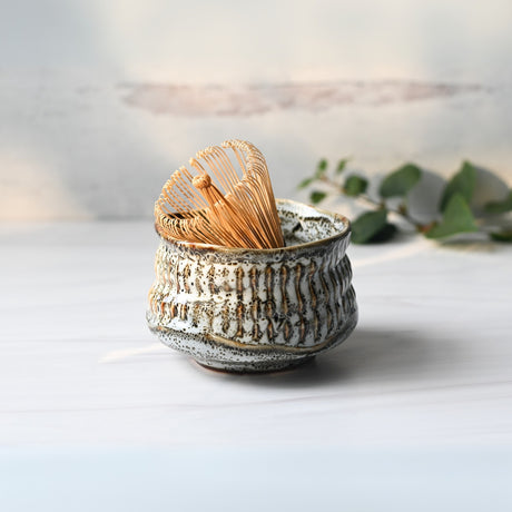 The White Ceramic Meisen Matcha Bowl and Bamboo Matcha Whisk with holder are elegantly set on a white surface, complemented by delicate eucalyptus leaves in the background.