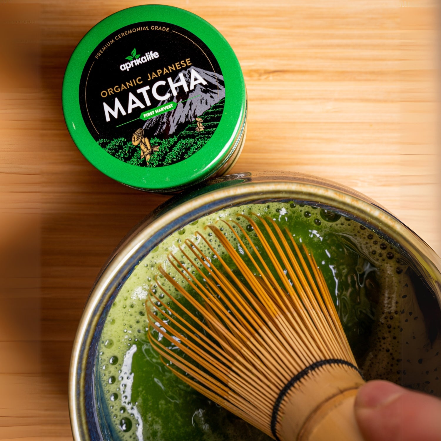 A bamboo whisk elegantly stirs organic matcha in a bowl, while a container of Organic Japanese Ceremonial Grade Matcha Green Tea Powder sits on the wooden surface in the background.