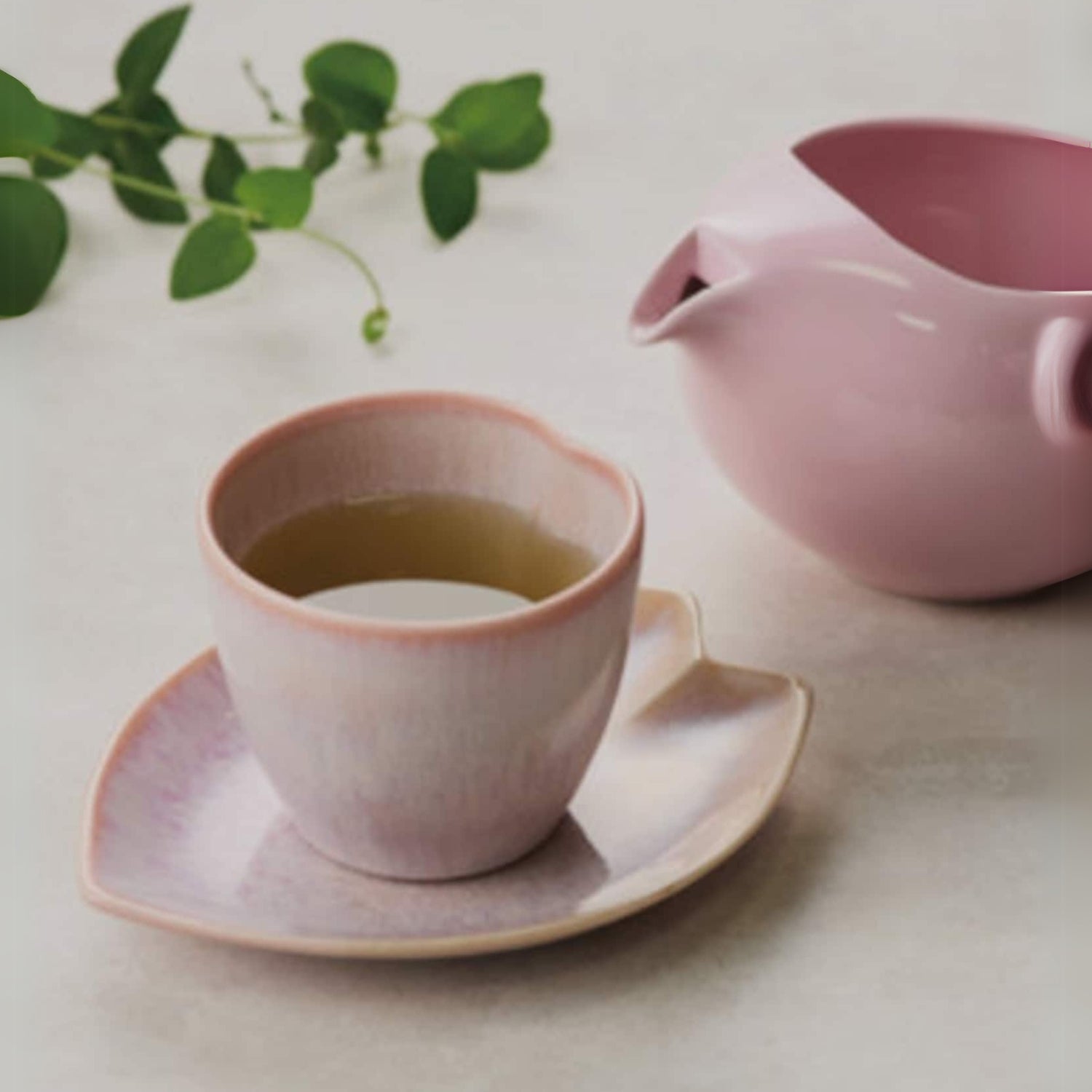 A Pink LOVE Ceramic Tea Cup with Saucer, filled with tea, sits beside a green leafy sprig on a light surface.