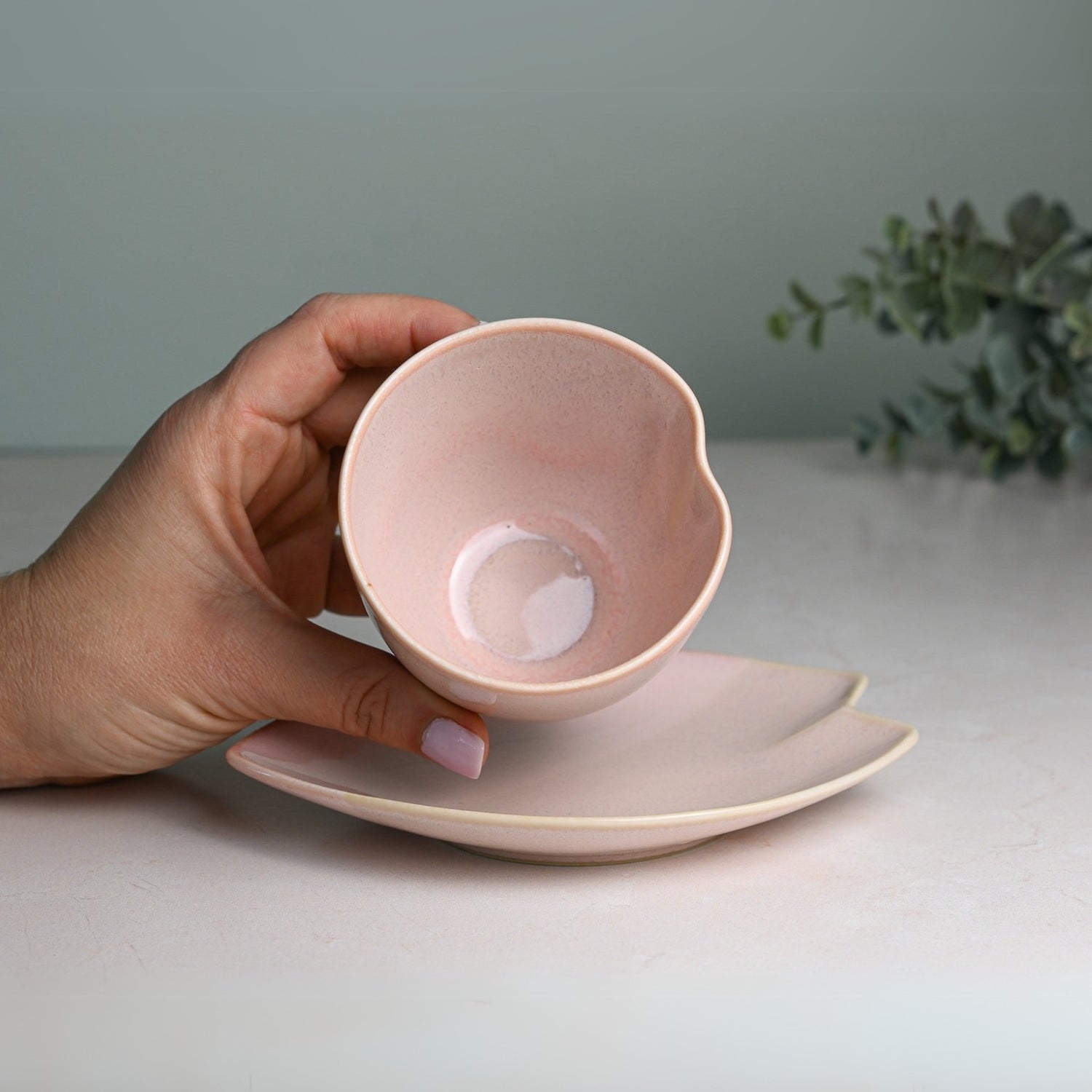 A hand gently holds a Pink LOVE Ceramic Tea Cup with a handle, floating above its matching saucer on a light surface.