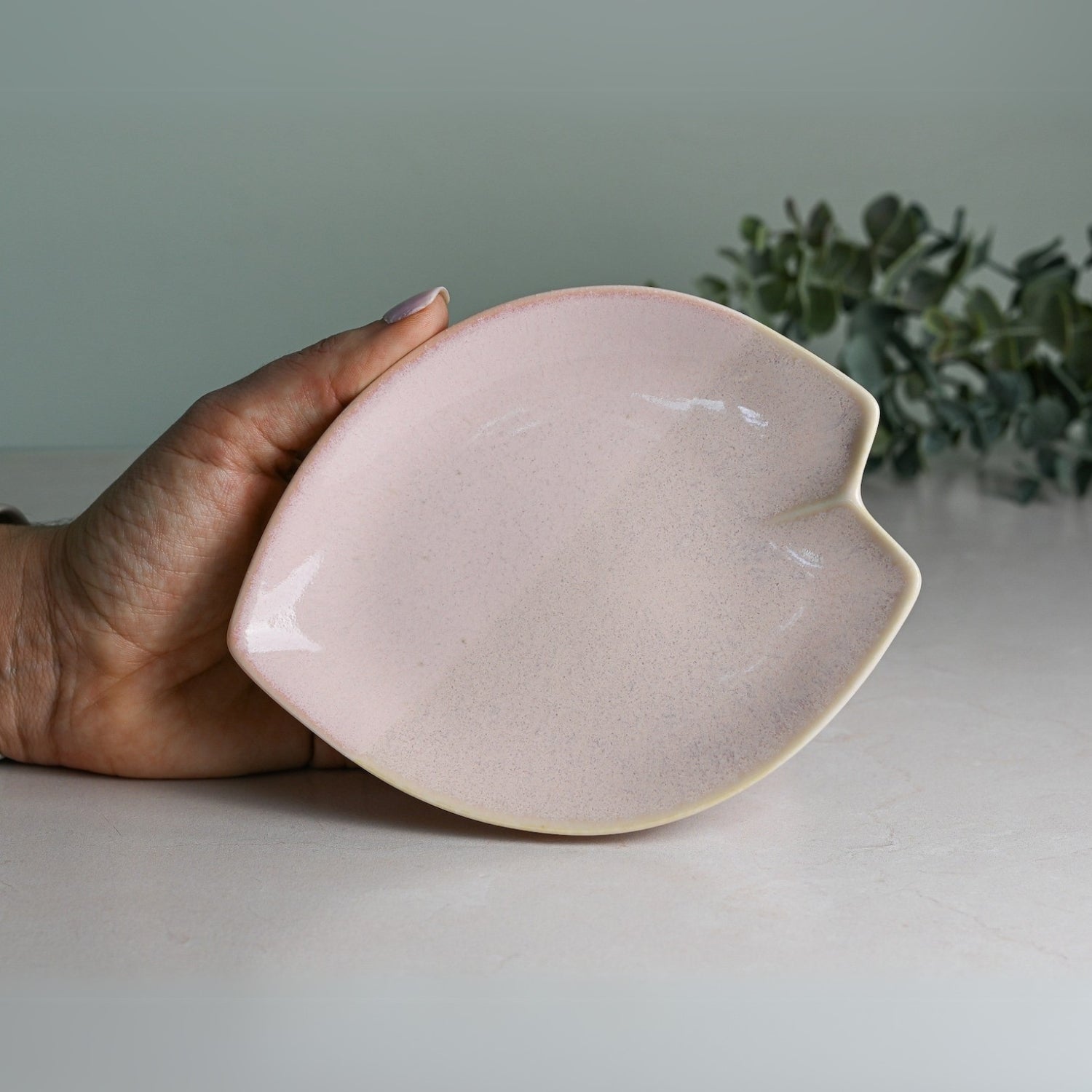 A hand holds a pink, leaf-shaped ceramic dish against green foliage, reminiscent of a Pink LOVE Ceramic Tea Cup with Saucer.