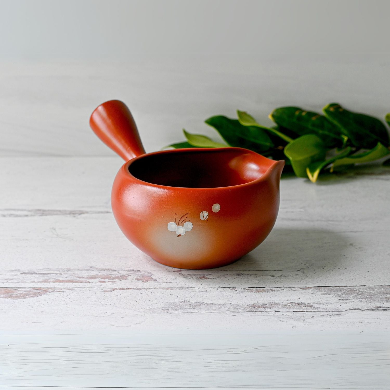 A brown teapot with a side handle and delicate floral designs sits next to a Red Flowers Ceramic Matcha Bowl with Spout and an Organic Japanese Matcha Set on a white wooden surface.