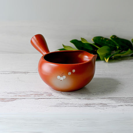 A brown teapot with a side handle and delicate floral designs sits next to a Red Flowers Ceramic Matcha Bowl with Spout and an Organic Japanese Matcha Set on a white wooden surface.