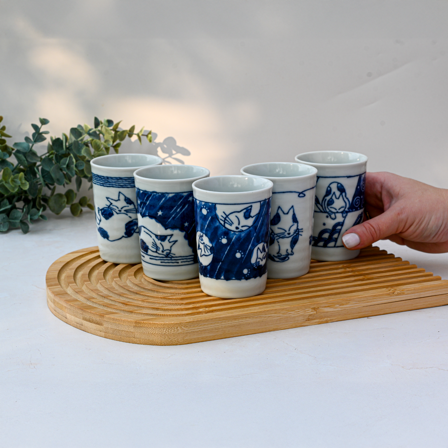 A hand holds one from a Set of 5 Adorable Cat Tea Cups, Blue & White Mino-Ware, 180ml (6oz.), while the other charming Japanese-crafted cups with blue cat designs rest on a wooden tray.