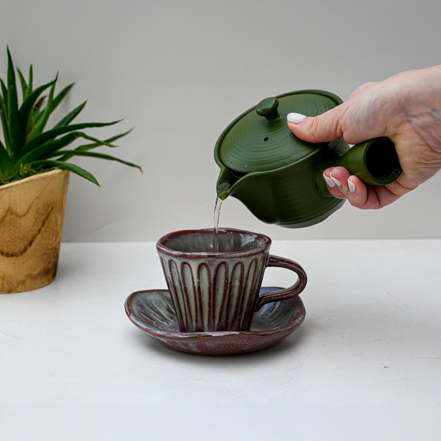 A hand pours tea from a green teapot into the Shibu Yunomi Japanese Tea Cup with Saucer, highlighting its handmade charm, with a potted plant nearby.
