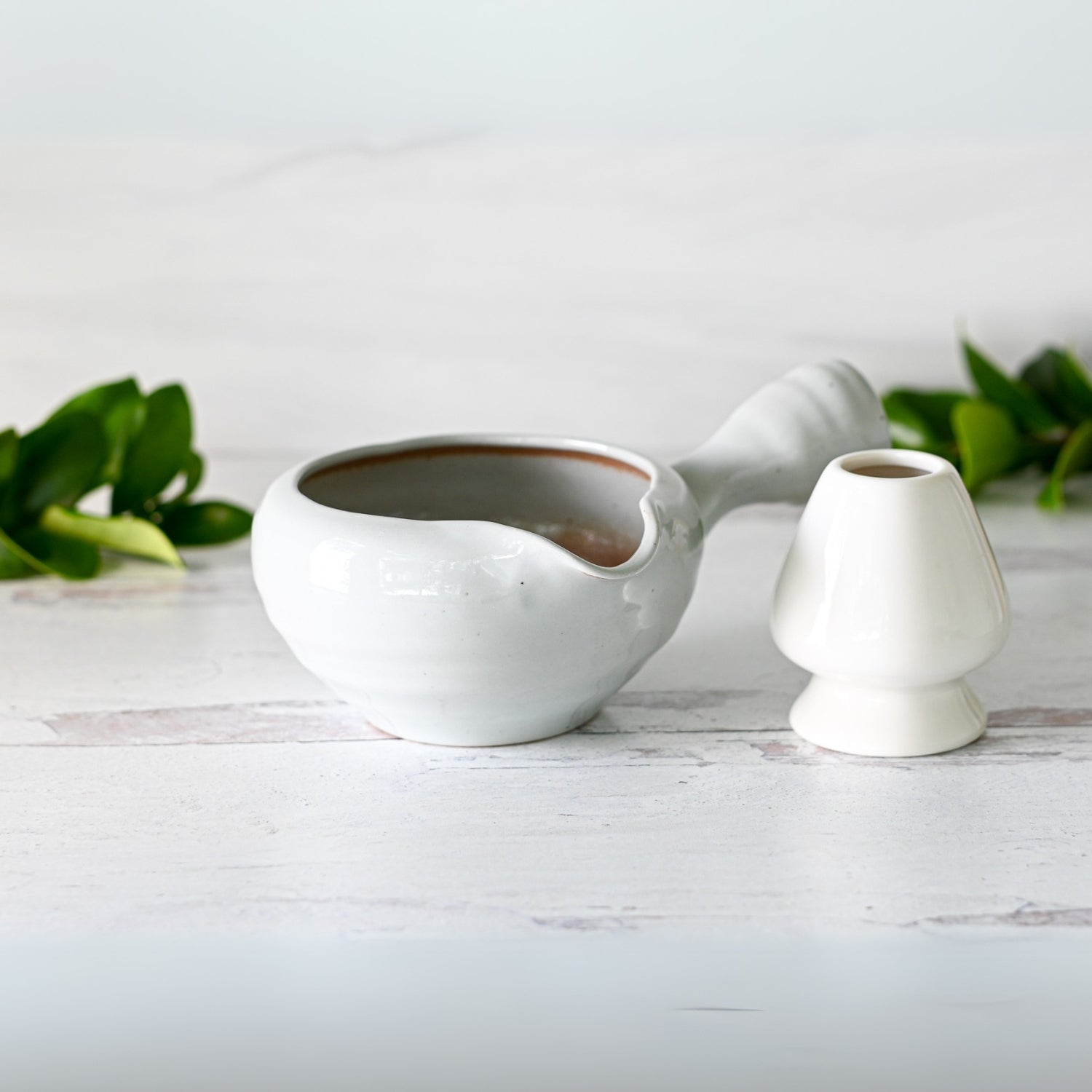 A gray spouted ceramic matcha bowl with a textured handle rests on a light wooden surface, accompanied by green leaves, bamboo matcha whisk, whisk holder, and organic Japanese matcha set, encapsulating the serene beauty of traditional Minoyaki craftsmanship.