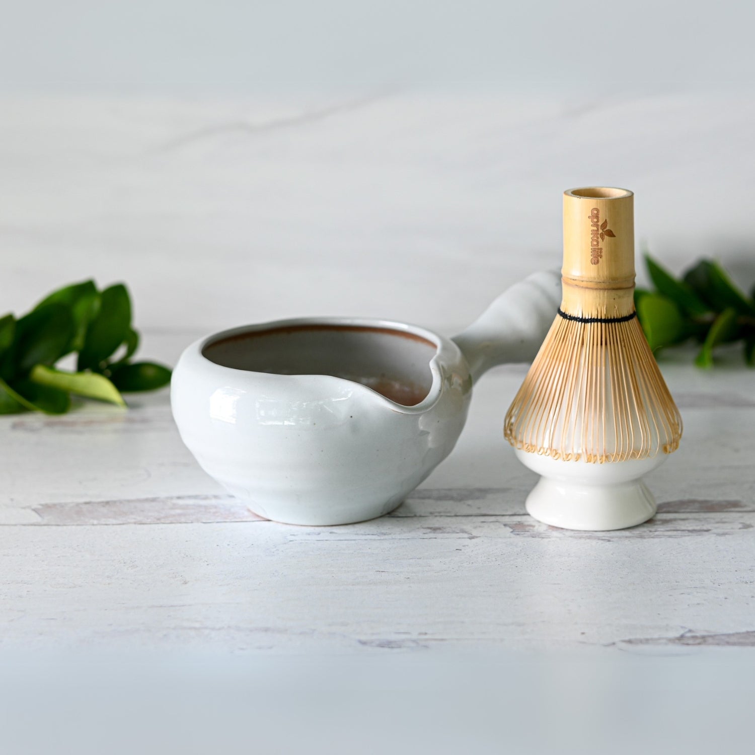The Gray Spouted Ceramic Matcha Bowl with Textured Handle, paired with a Bamboo Matcha Whisk and Holder, is elegantly displayed on a light wooden surface amidst green leaves, capturing the essence of traditional Japanese craftsmanship.