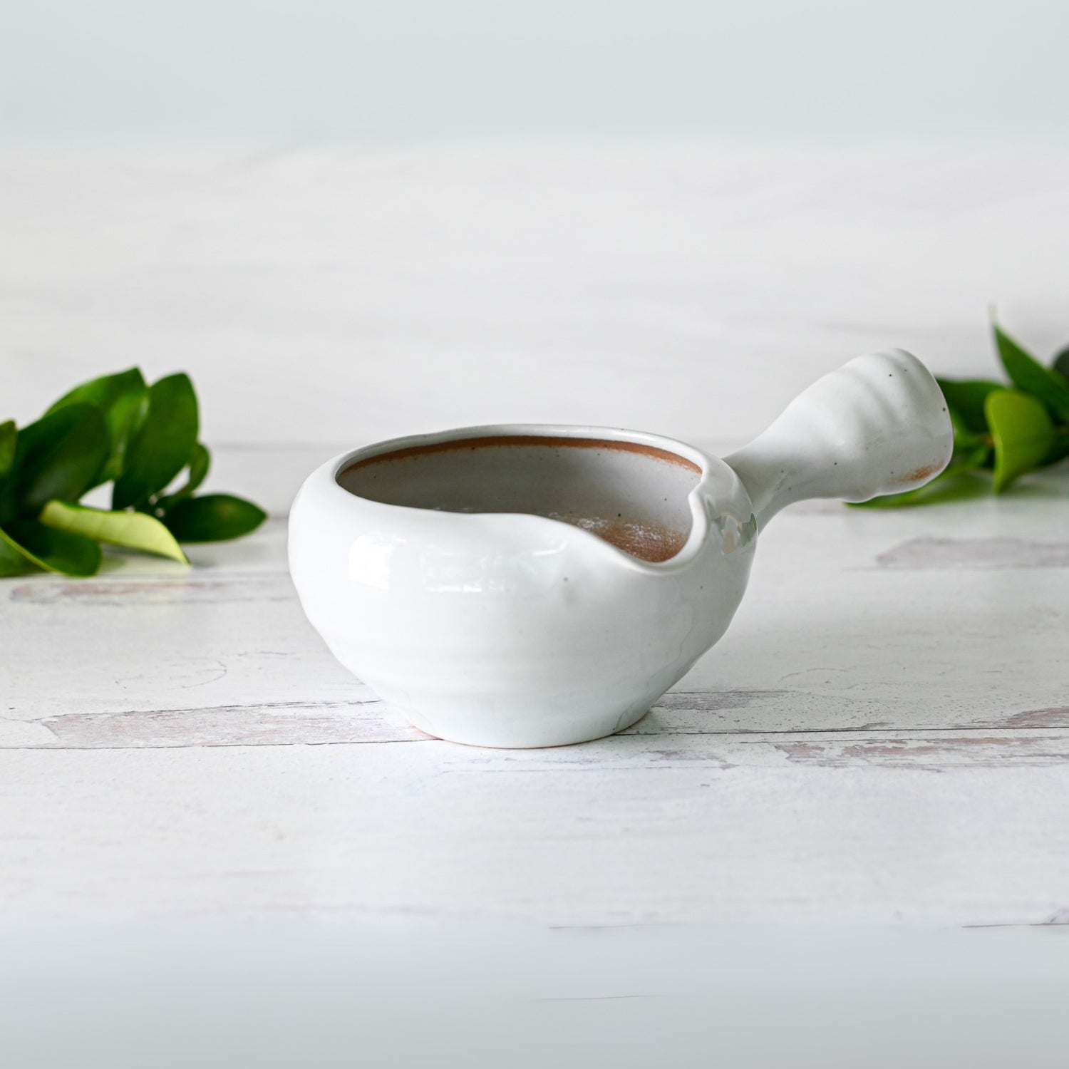 A gray spouted ceramic matcha bowl with a textured handle sits on a light wooden surface, surrounded by lush green leaves, evoking the essence of Japanese pottery.