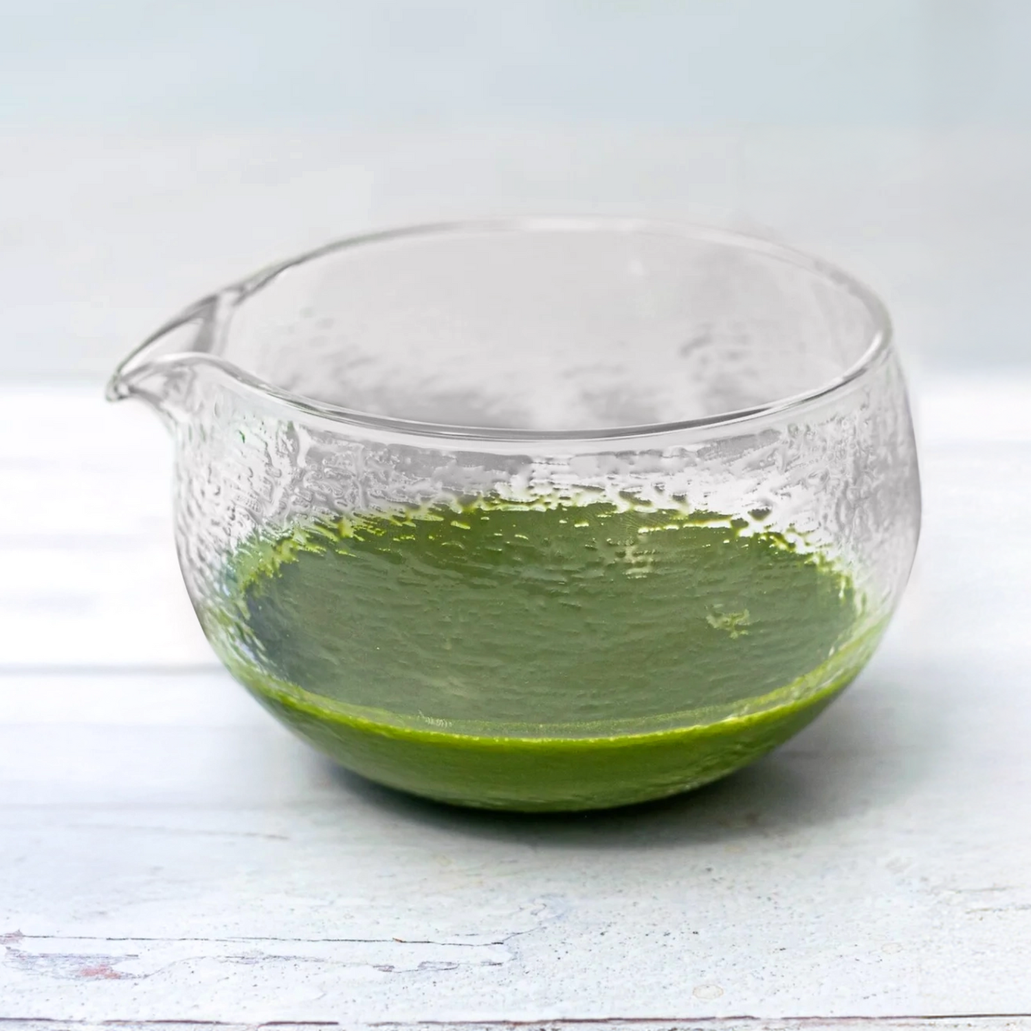 The Textured Spouted Glass Matcha Bowl Pattern 1, holding a small amount of green matcha, sits on a light wooden surface, showcasing minimalist teaware elegance.