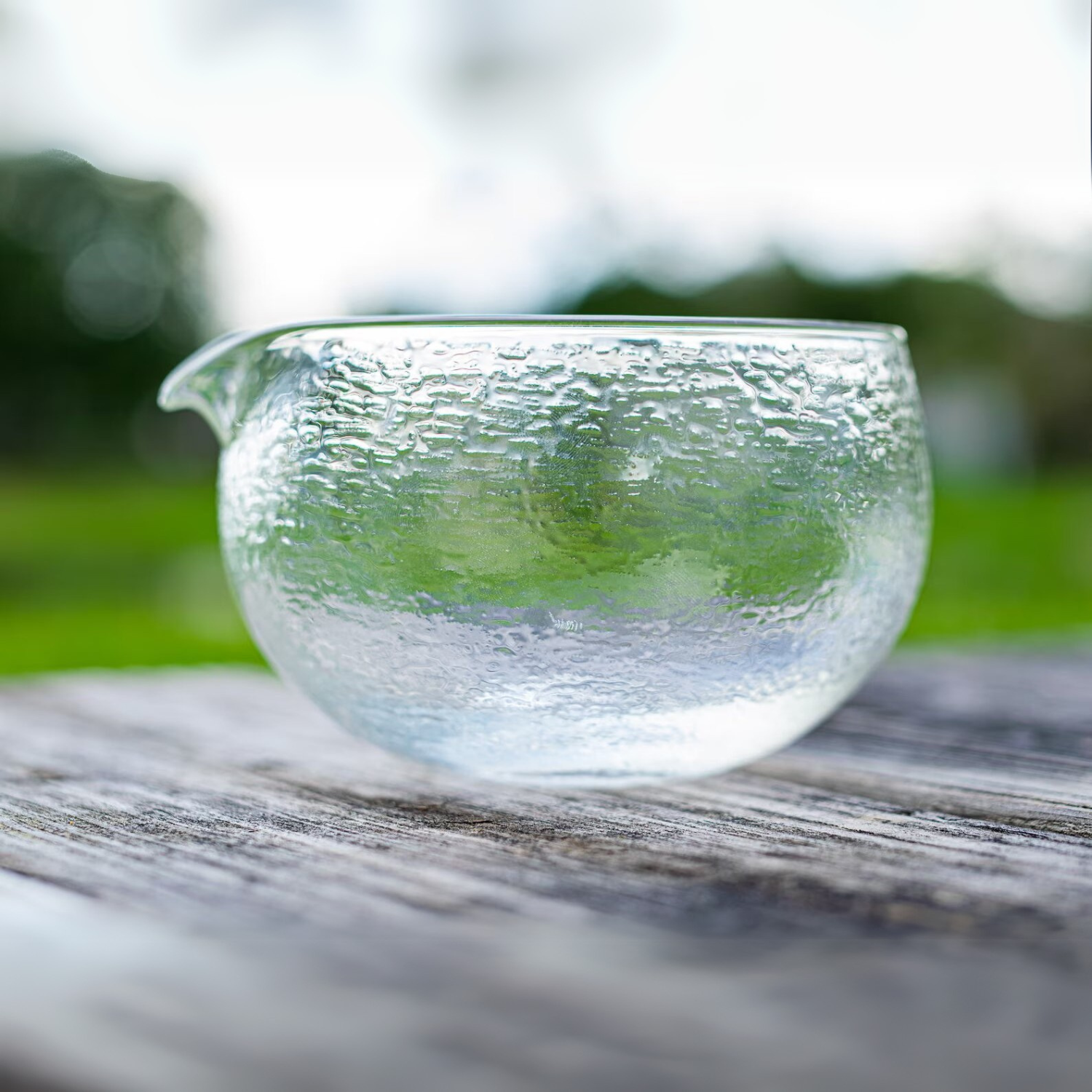 The Textured Spouted Glass Matcha Bowl Pattern 1 sits on a wooden surface outdoors, surrounded by lush greenery.