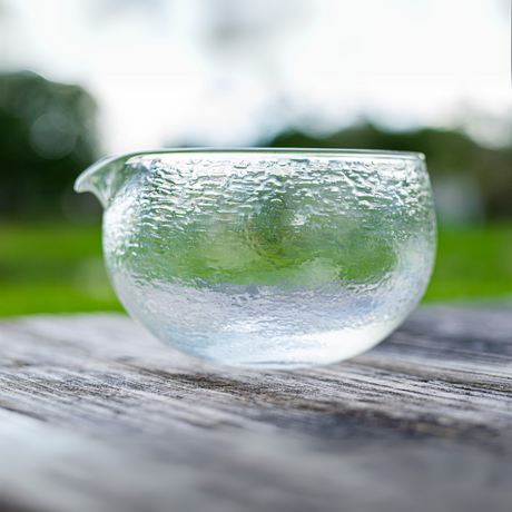 The Textured Spouted Glass Matcha Bowl Pattern 1 with bamboo whisk and holder set rests on a wooden surface, perfect for your matcha ritual; blurred greenery adds to the serene background.