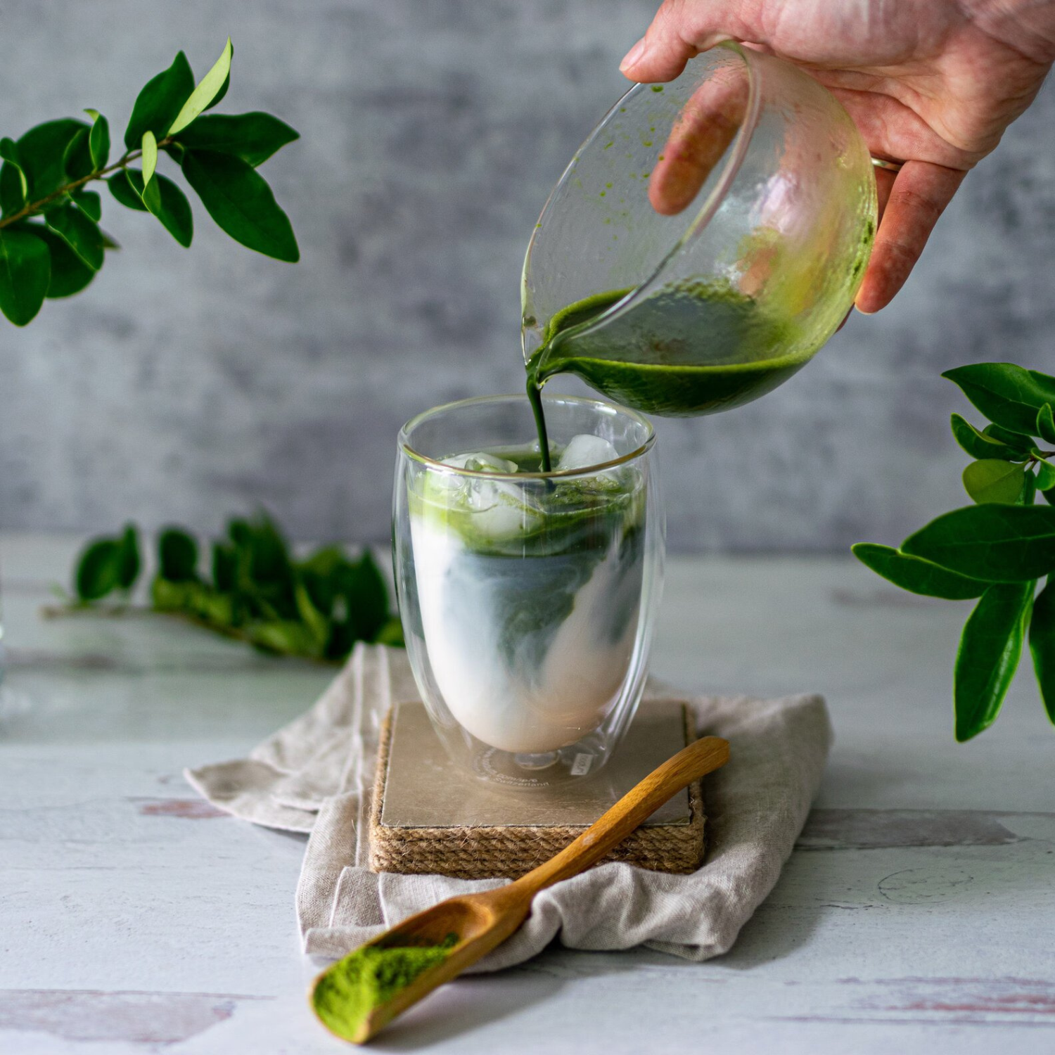 A hand pours green matcha from the Textured Spouted Glass Matcha Bowl Pattern 1 into iced milk, with the Bamboo Matcha Whisk and Whisk Holder Set displayed nearby.