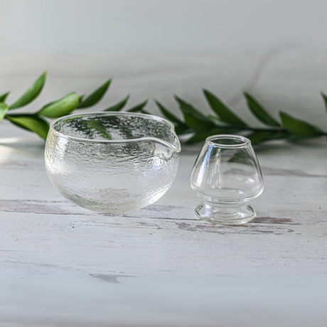 A clear glass pitcher and small cup sit on a white surface with green leaves, perfectly complemented by the Textured Spouted Glass Matcha Bowl Pattern 1 from the Bamboo Matcha Whisk and Whisk Holder Set.