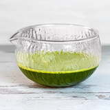 The Textured Spouted Glass Matcha Bowl Pattern 2, filled halfway with vibrant green matcha, rests elegantly on a light wooden surface.