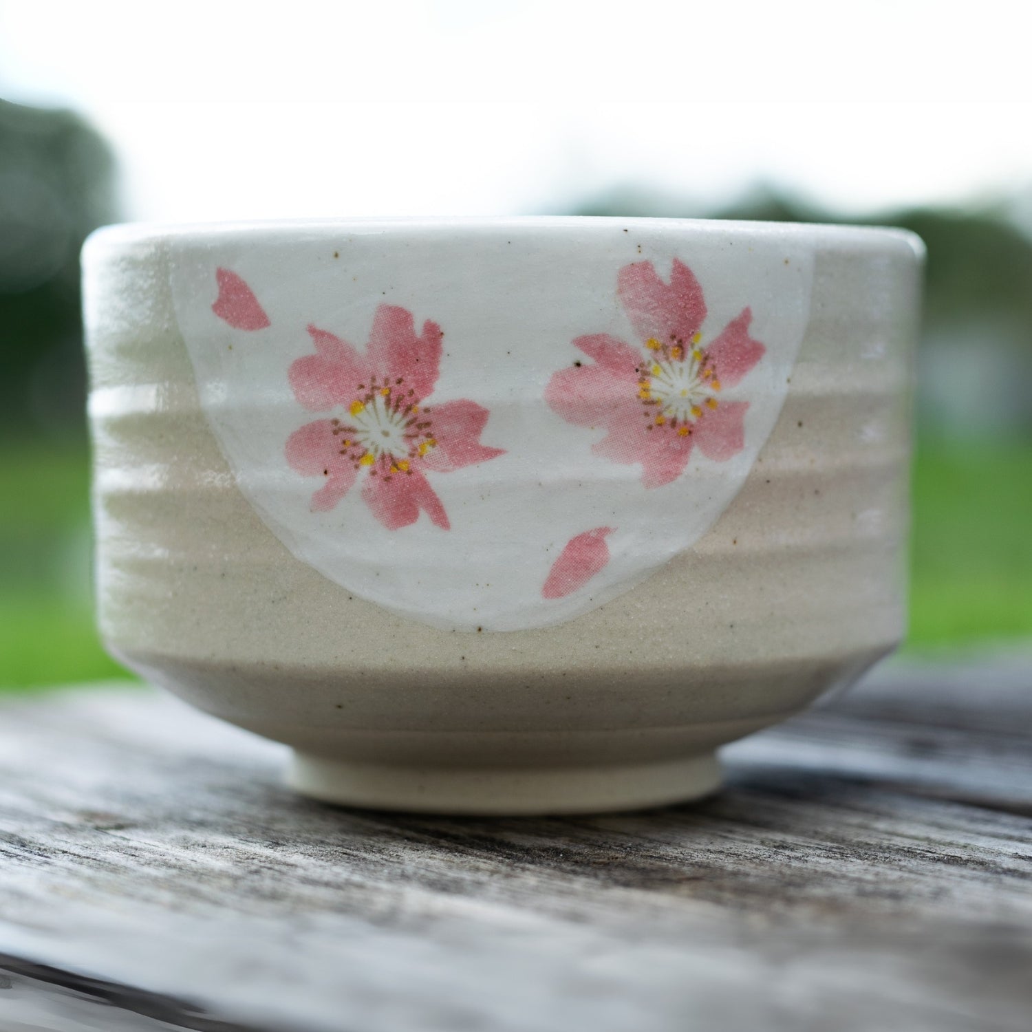 The Beige with Pink Flowers Matcha Bowl, Bamboo Matcha Whisk and Whisk Holder Set is displayed on a wooden surface outdoors, its elegant floral design inspired by traditional artistry.