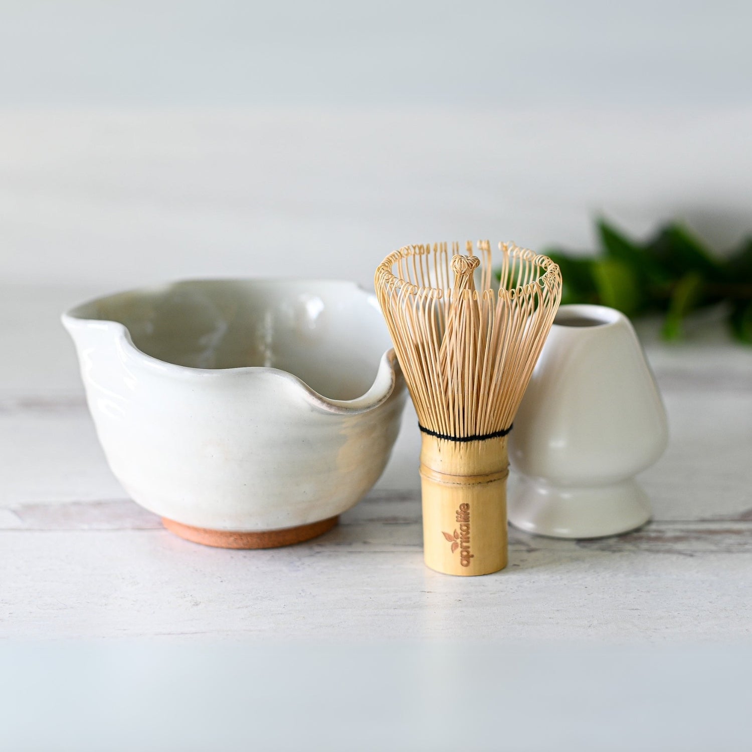 White wavy ceramic matcha bowl with spout paired with a bamboo whisk on a white wooden surface.