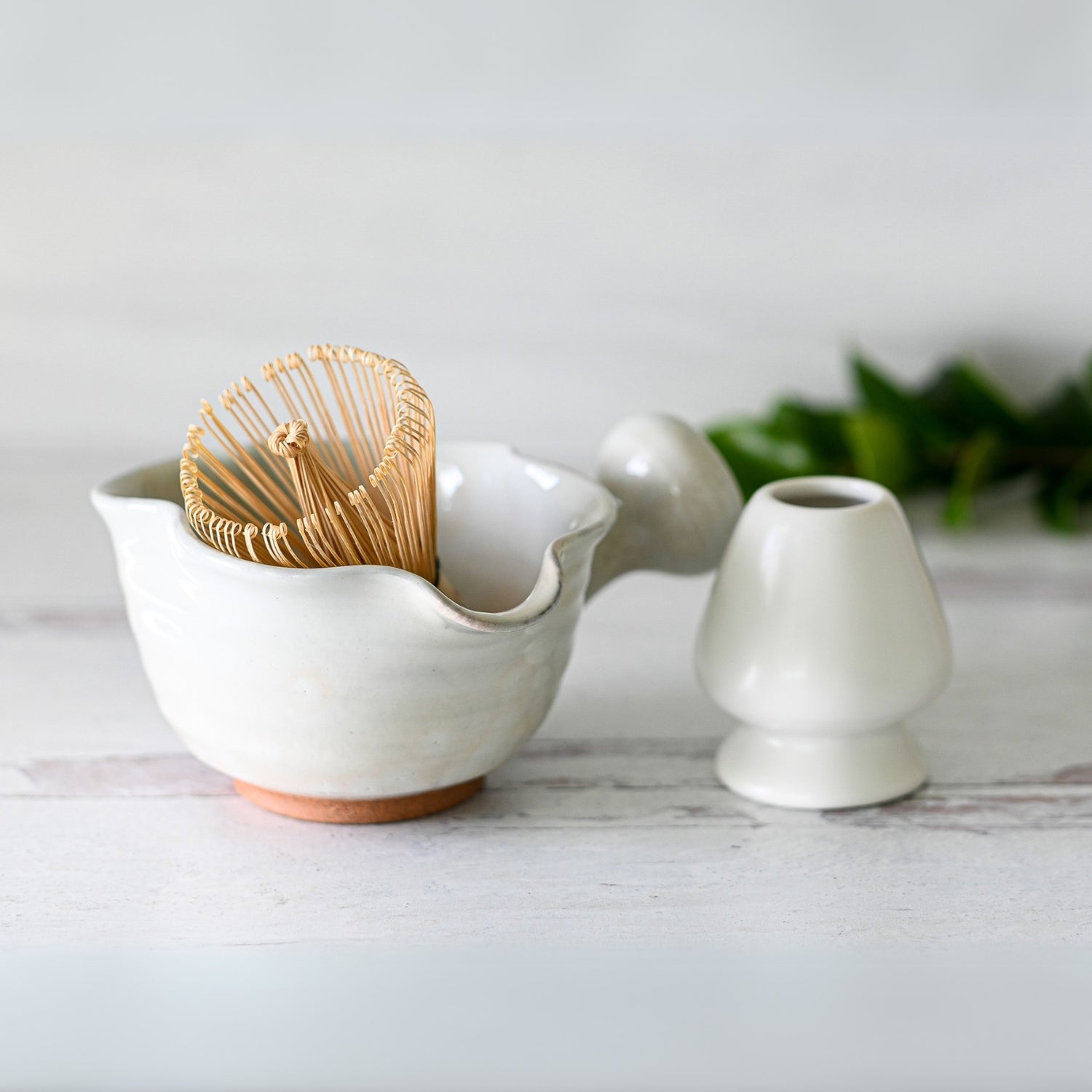 A White Wavy Ceramic Matcha Bowl with Spout and optional bamboo whisk set rests on a light surface, blurred greenery in the background.