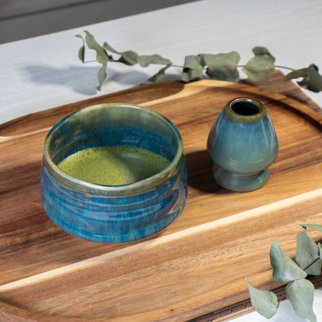 The Ceremonial Matcha Starter Kit - Blue includes a ceramic bowl with ceremonial matcha tea and a small vase on a wooden tray, inspired by Japanese tea ceremonies and adorned with eucalyptus leaves.